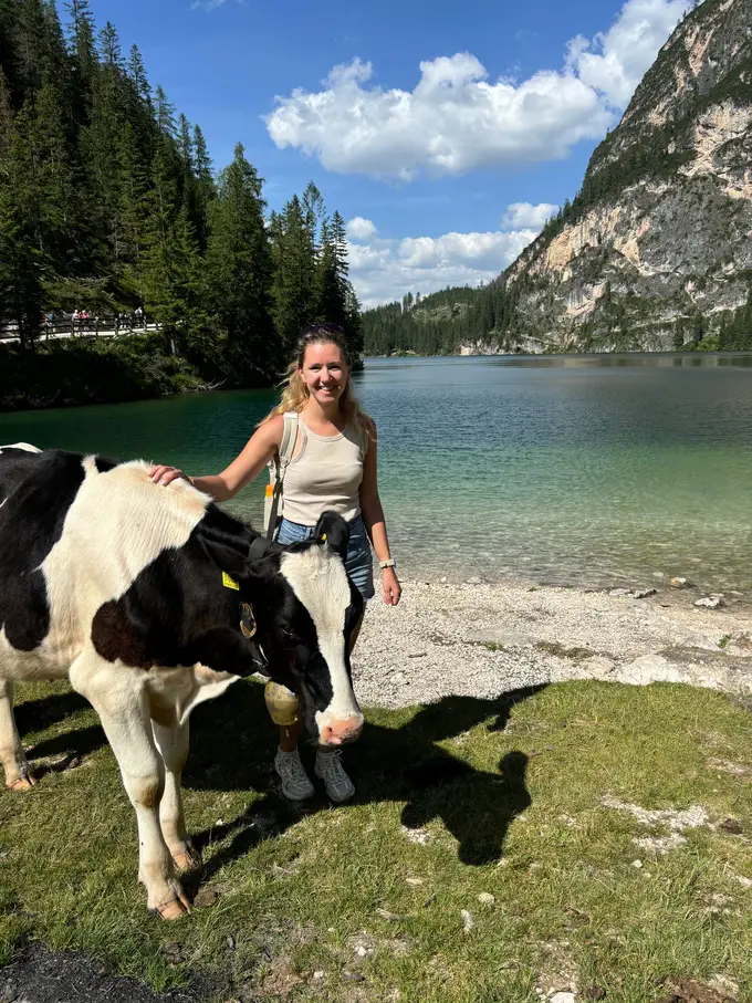 Rebecca legt lächelnd die Hand auf eine schwarz-weiße Kuh mit Glocke, direkt dahinter das Kiesufer und der klare, grüne See