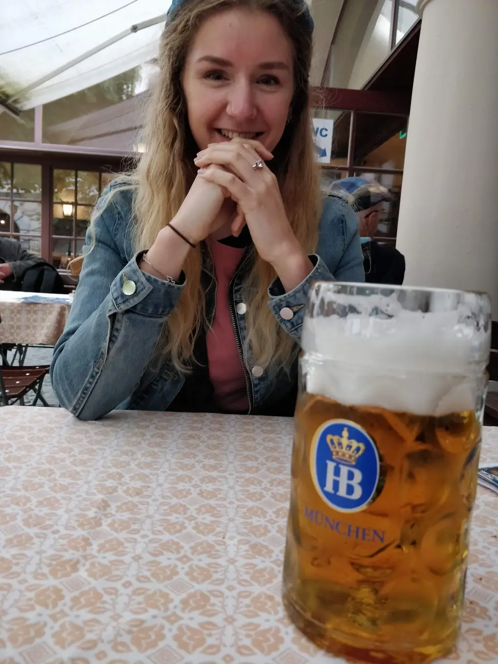 Rebecca sitzt lächelnd an einem Tisch mit gemusterter Tischdecke, vorne eine volle Maß mit dem Logo des Hofbräuhauses