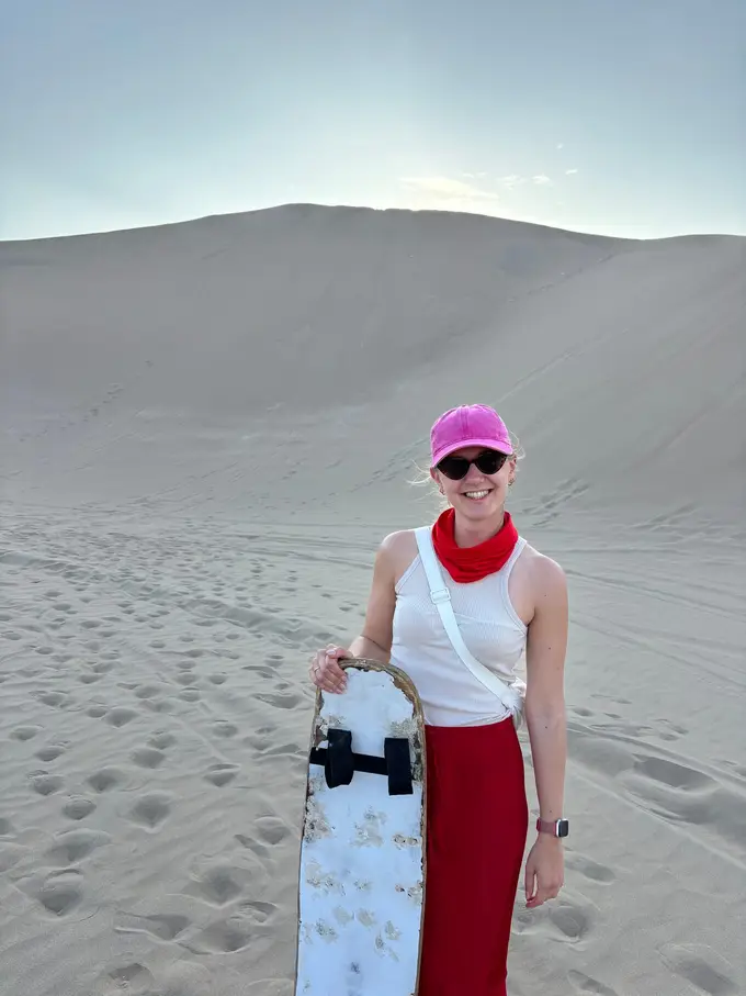 Rebecca mit pinker Kappe und rotem Halstuch hält ein Sandboard, hinter ihr ein steiler Dünenhang voller Fußspuren