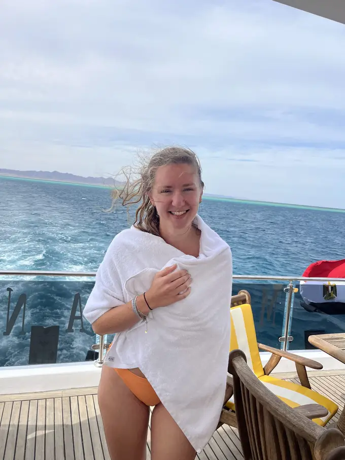 Rebecca steht mit nassen Haaren und in ein Handtuch gewickelt an Deck, hinter ihr das Heckwasser und eine ägyptische Flagge