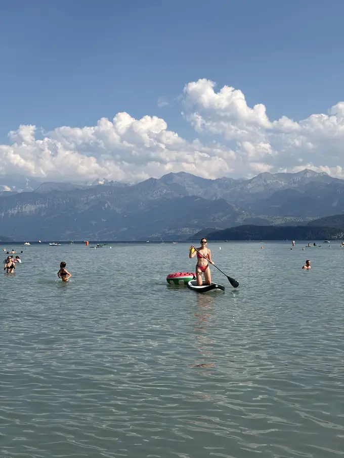 Rebecca kniet mit Paddel auf dem Board im flachen Wasser, daneben der Schwimmring, im Hintergrund Badende und die Berge auf der anderen Seeseite