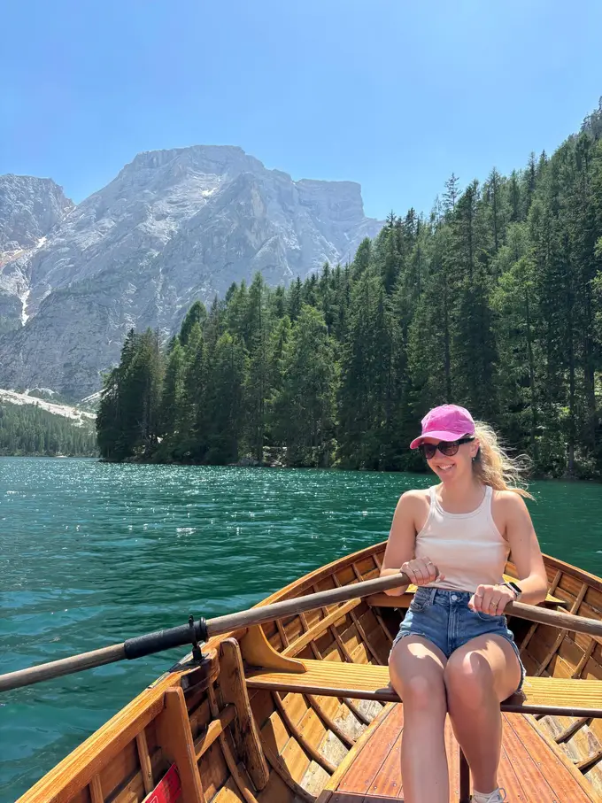 Rebecca mit pinker Kappe rudert lachend im Holzboot, hinter ihr grünes Wasser, ein Nadelwald am Ufer und ein Felsberg