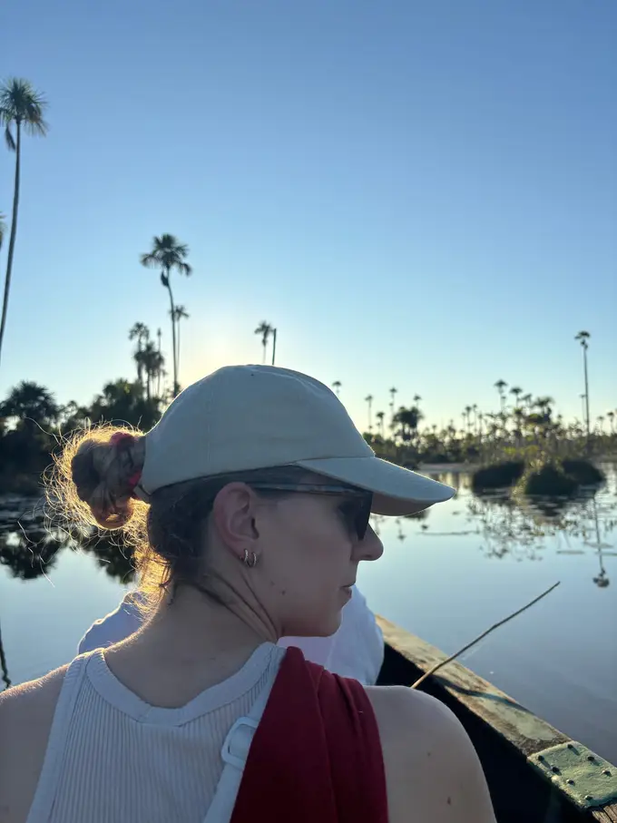 Rebecca mit Schirmmütze und Sonnenbrille sitzt im Holzboot und blickt über den See, im Hintergrund Palmen und die tiefstehende Sonne