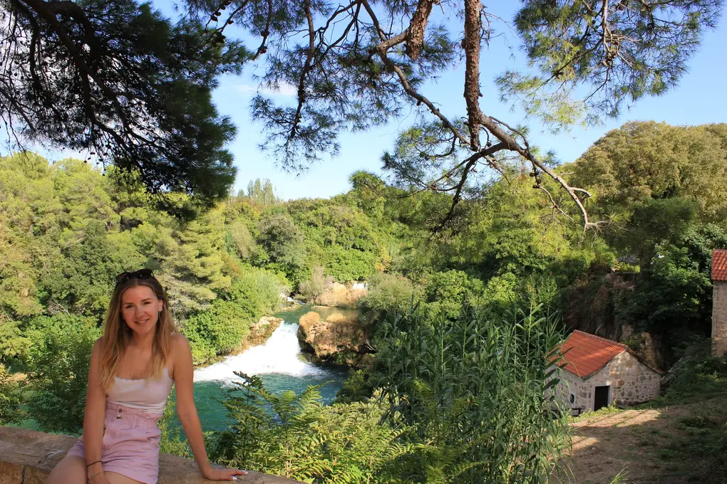 Rebecca sitzt auf einer Steinmauer unter einer Kiefer, unten fällt ein kleiner Wasserfall in ein türkises Becken, daneben ein Steinhäuschen mit rotem Dach
