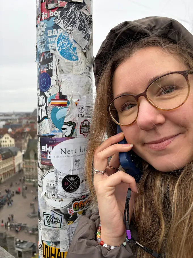 Rebecca mit Brille und Kapuze lächelt mit dem Handy am Ohr neben einem mit Aufklebern übersäten Mast, tief unter ihr ein Platz und die Dächer der Genter Altstadt
