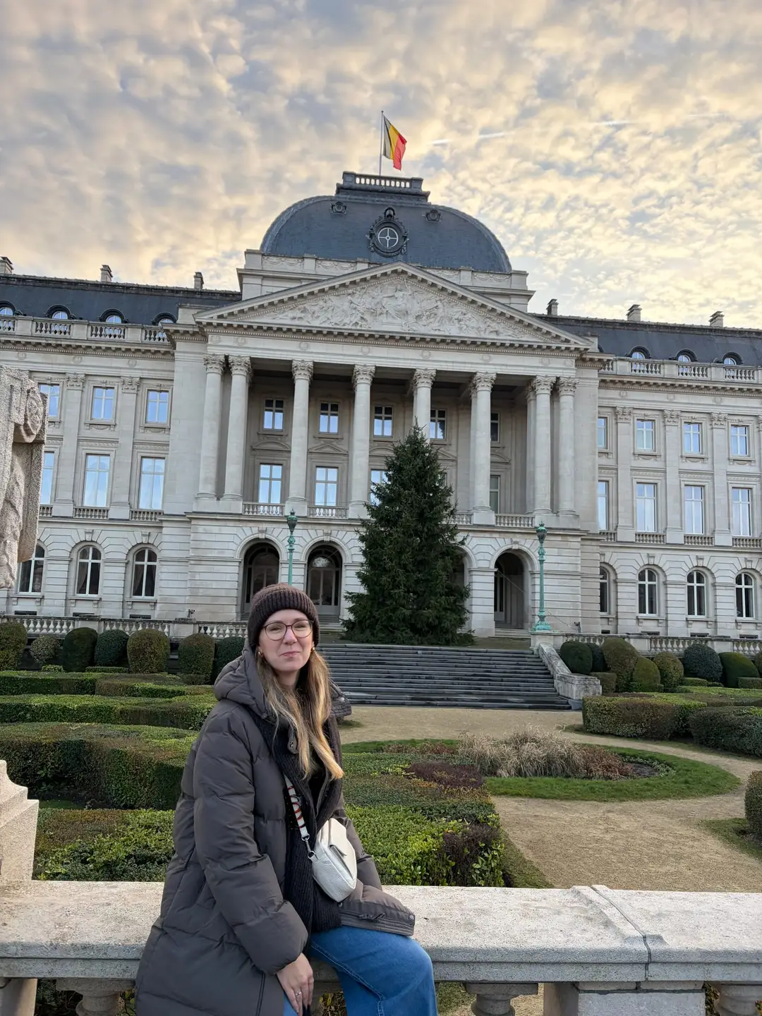 Rebecca sitzt auf einer steinernen Brüstung, hinter ihr ein Garten mit Hecken und der Königspalast mit Säulenportal, Kuppel und belgischer Flagge