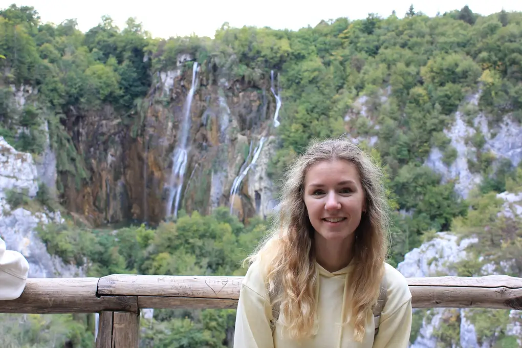 Rebecca an einem Holzgeländer, hinter ihr eine hohe Felswand, über die mehrere Wasserstränge fallen
