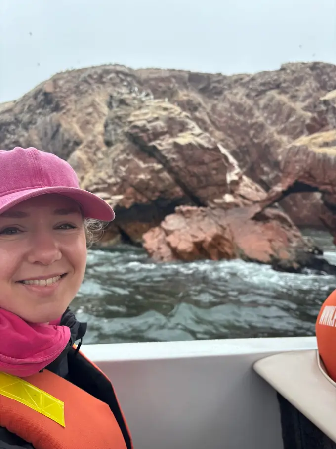 Rebecca mit pinker Kappe und Schwimmweste lächelt im Boot, hinter ihr zerklüftete rötliche Felsen der Inseln