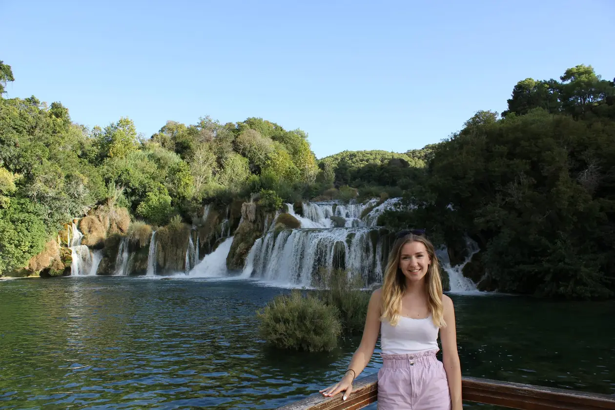 Rebecca lehnt an einem Holzgeländer, hinter ihr fällt das Wasser in mehreren breiten Stufen in ein grünes Becken