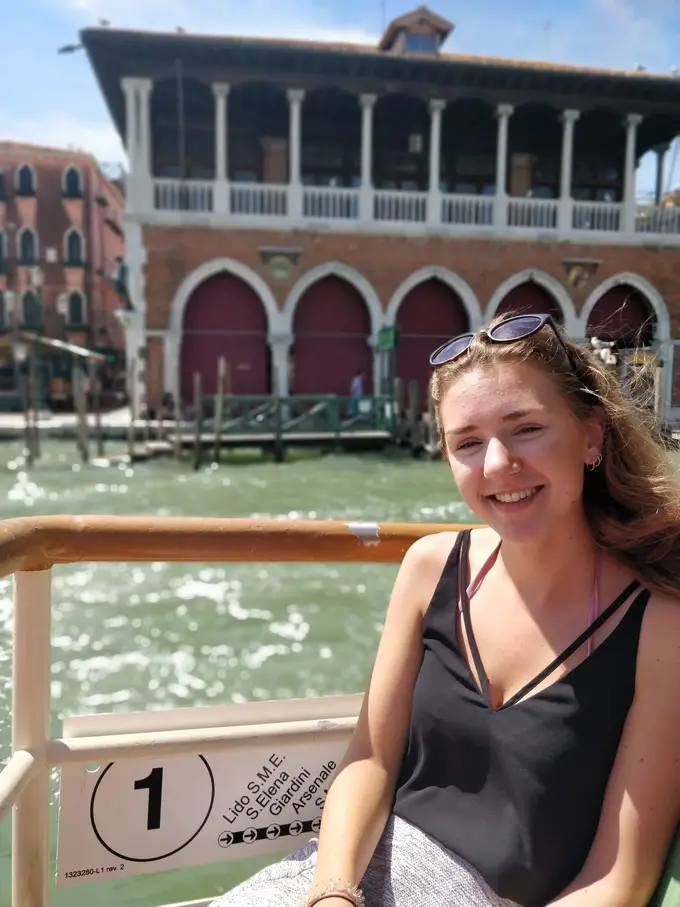 Rebecca an der Reling eines Boots, hinter ihr die Markthalle mit roten Bögen am Canal Grande