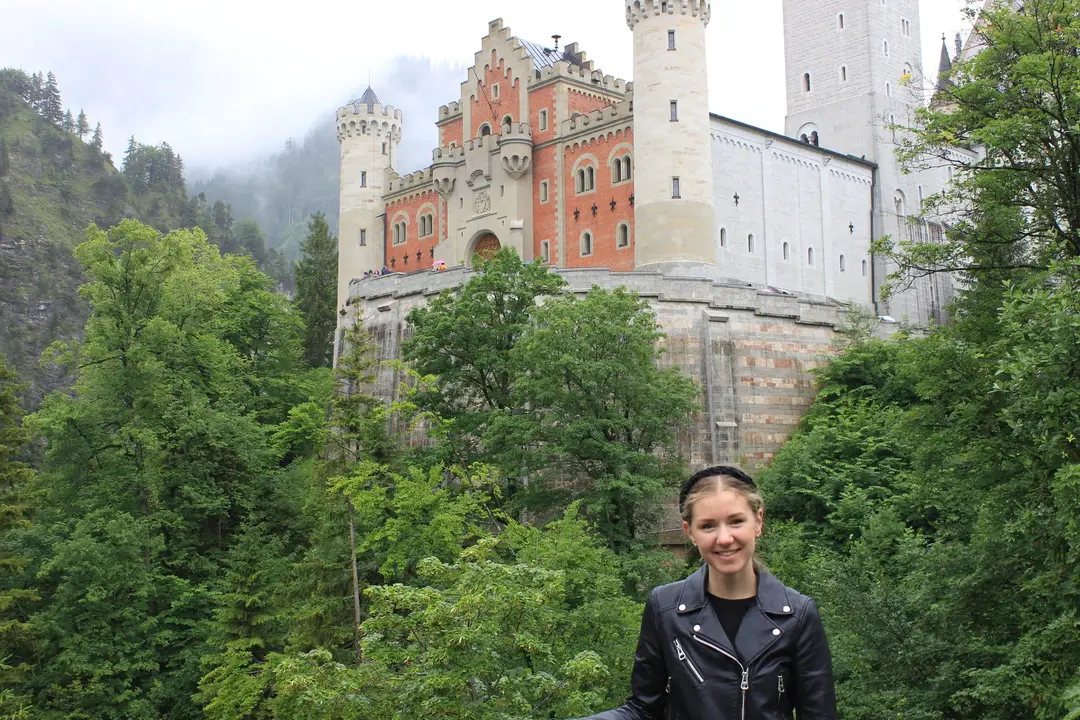 Rebecca steht vor einem bewaldeten Hang, hinter ihr Schloss Neuschwanstein mit hellen Rundtürmen, roter Backsteinfassade und hoher Mauer, der Berg dahinter im Nebel