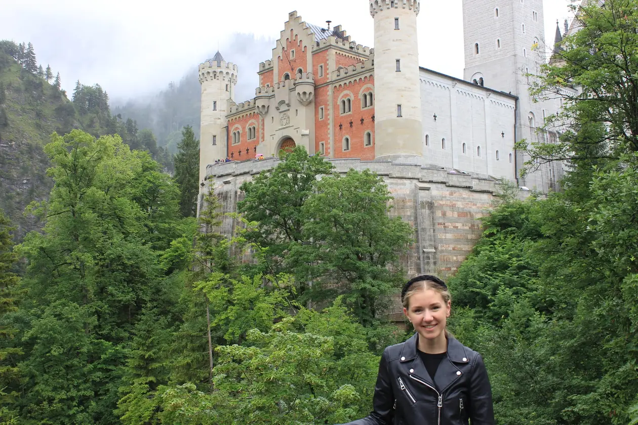 Rebecca steht vor einem bewaldeten Hang, hinter ihr Schloss Neuschwanstein mit hellen Rundtürmen, roter Backsteinfassade und hoher Mauer, der Berg dahinter im Nebel