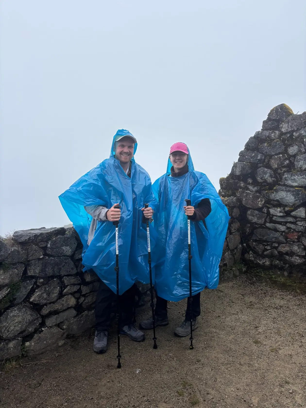 Max und Rebecca in blauen Regenponchos zwischen Inka-Mauern aus grauem Stein, dahinter nur Nebel