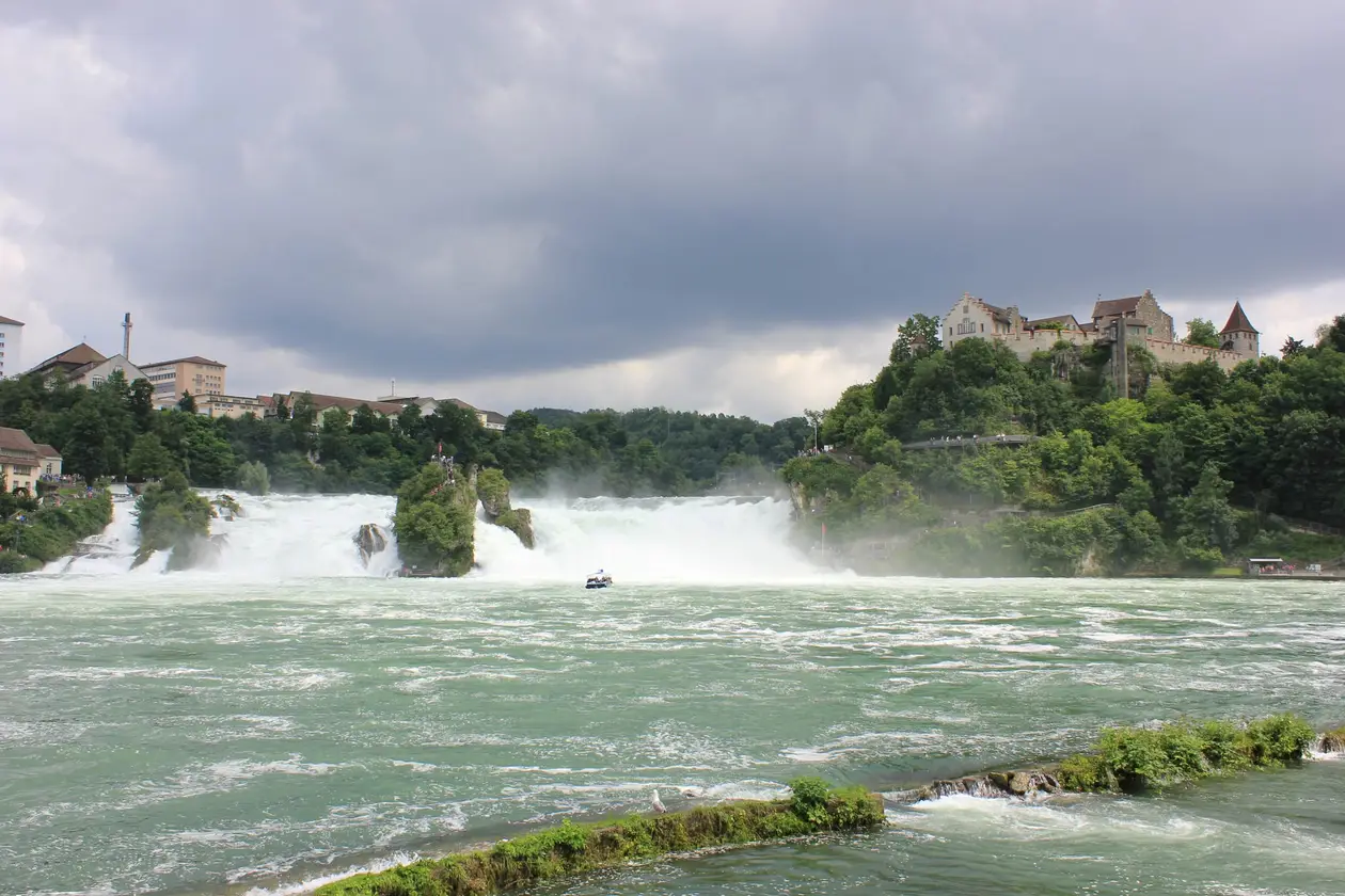 Der Rheinfall unter dunklen Wolken, weißes Wasser stürzt über die Felsen, auf einem Felsen in der Mitte eine Fahne, rechts oben auf dem bewaldeten Hang ein Schloss, im Vordergrund aufgewühltes grünes Wasser