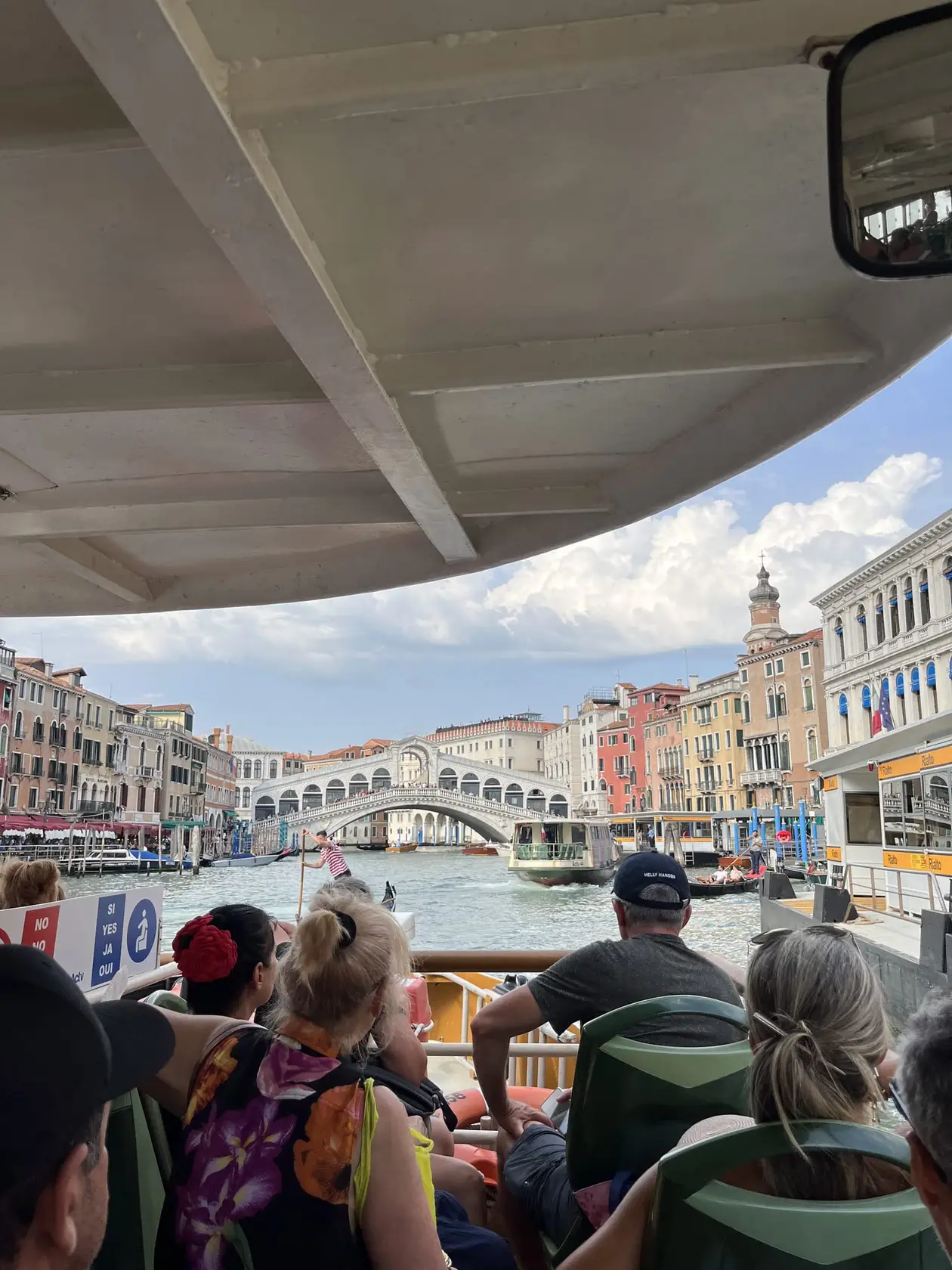 Blick unter dem Dach eines voll besetzten Vaporetto hindurch auf den Canal Grande mit der Rialtobrücke, davor ein Gondoliere und ein weiteres Linienboot