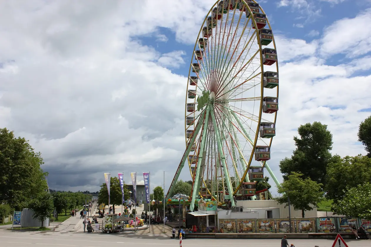 Ein großes Riesenrad mit bunten Gondeln unter Wolken, davor Fahnen, ein Kinderkarussell und Buden an einer Straße