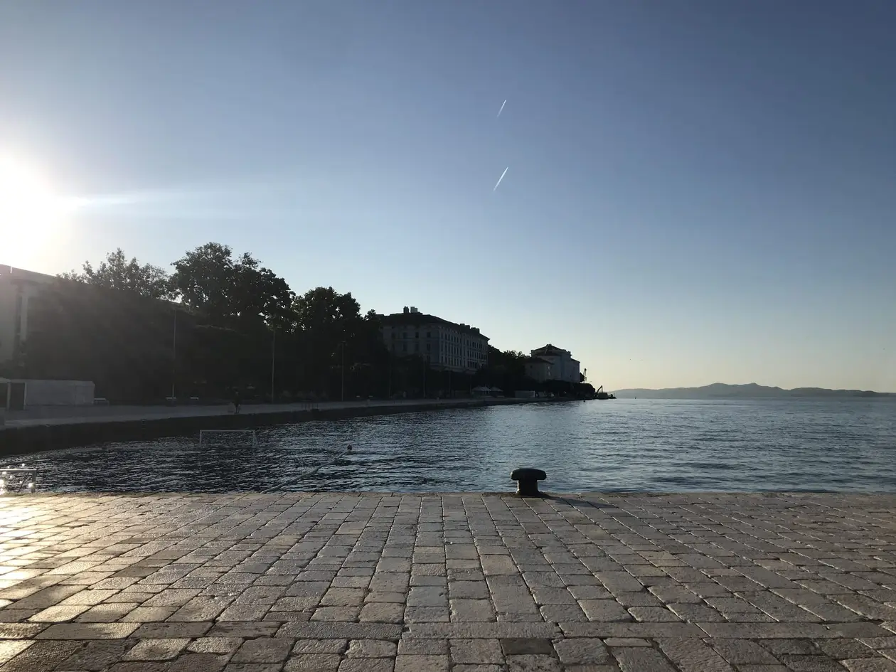Ein Steinkai mit Poller am ruhigen Wasser, darin ein Wasserballtor, am Ufer gegenüber Häuser, links tief die Sonne