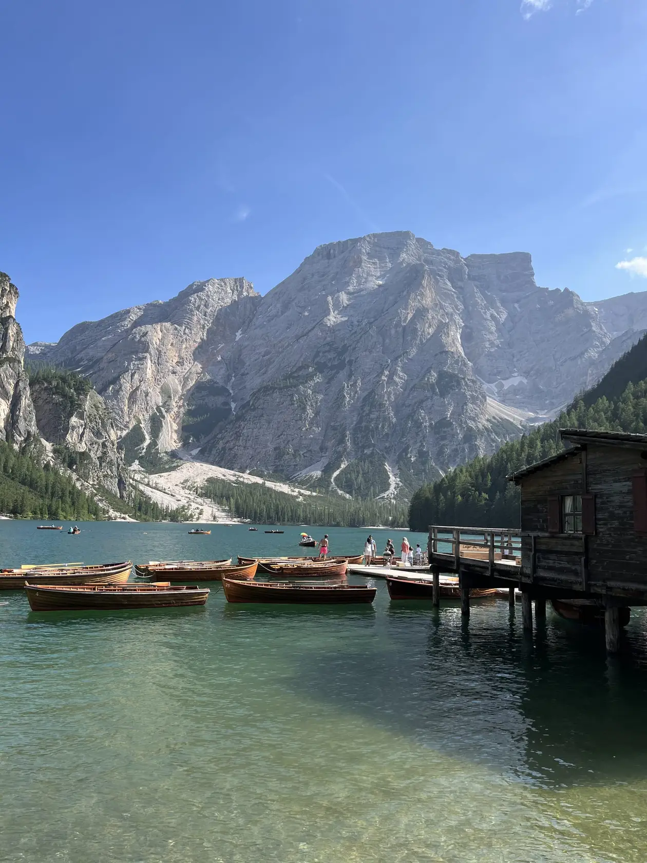 Hölzerne Ruderboote liegen vor einem dunklen Bootshaus auf Stelzen im grünen, klaren Wasser, dahinter ein großes helles Felsmassiv unter blauem Himmel