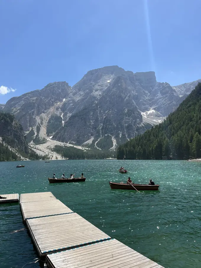 Ein Holzsteg ragt in den grünen See, auf dem Wasser mehrere Ruderboote mit Menschen, dahinter der Seekofel unter blauem Himmel