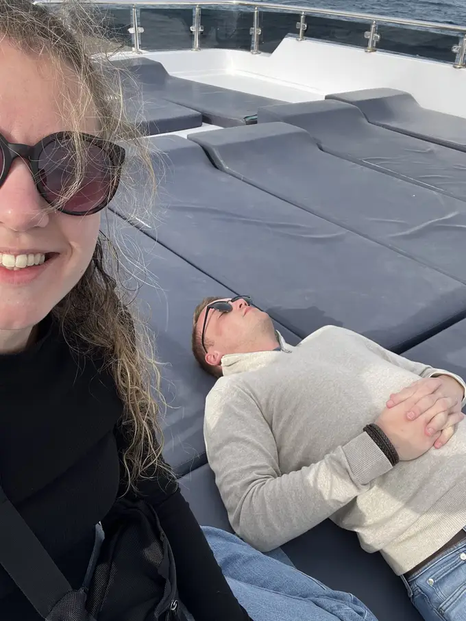 Selfie von Rebecca, hinter ihr liegt Max mit Sonnenbrille und geschlossenen Augen auf den Polstern an Deck