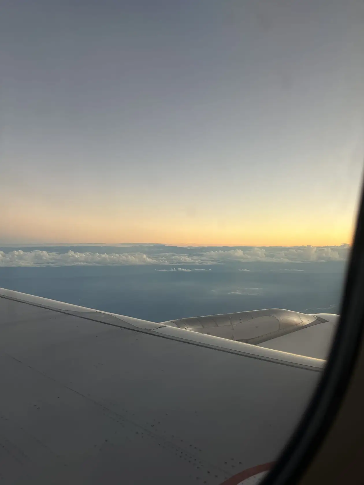 Blick aus dem Flugzeugfenster über die Tragfläche auf eine Wolkendecke, am Horizont ein gelber Streifen Abendlicht