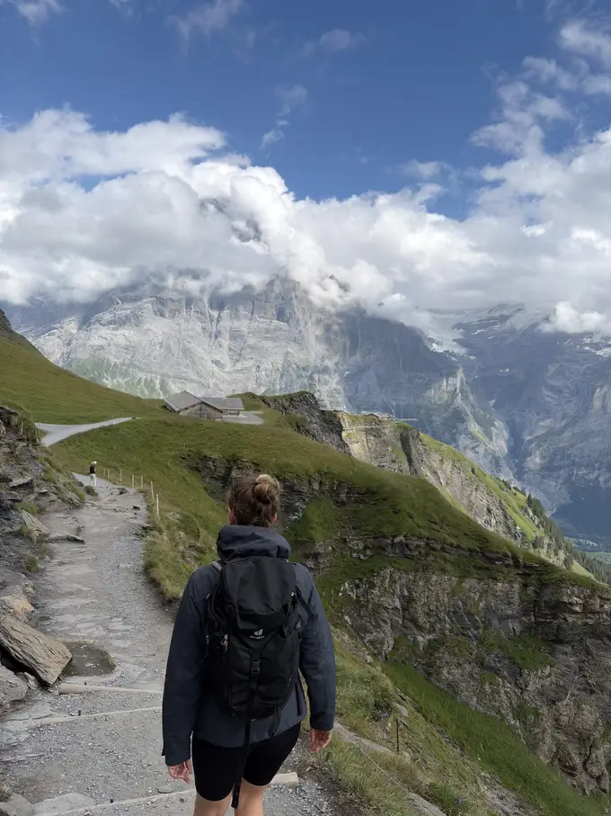 Rebecca geht von hinten gesehen auf einem steinigen Weg an einem Grashang entlang, vorne eine Hütte und dahinter mächtige Felswände in den Wolken
