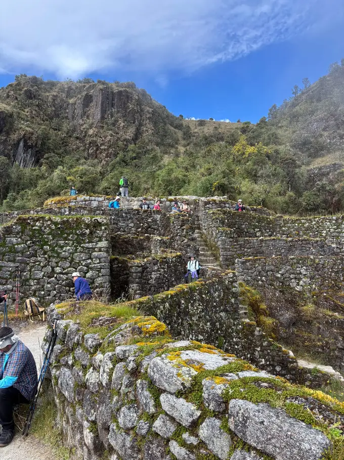 Inka-Anlage mit moosbewachsenen Steinmauern an einem Hang, oben auf den Mauern stehen Wanderer, dahinter Felsen und Nebel