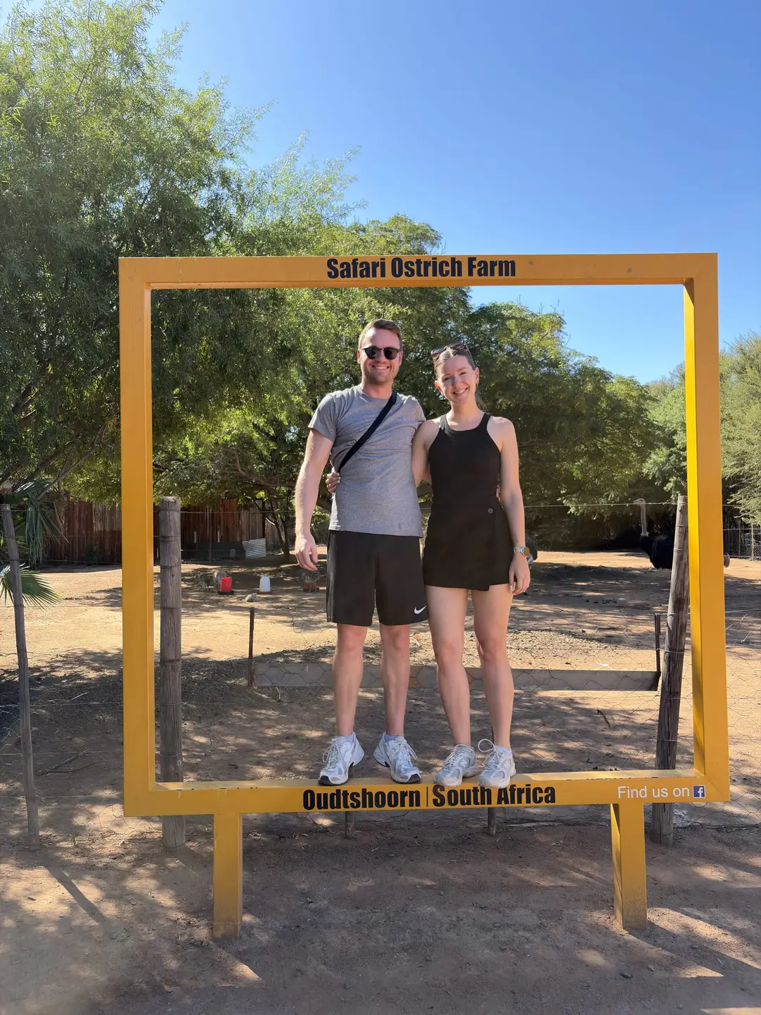 Max und Rebecca stehen in einem gelben Holzrahmen mit der Aufschrift Safari Ostrich Farm, Oudtshoorn South Africa
