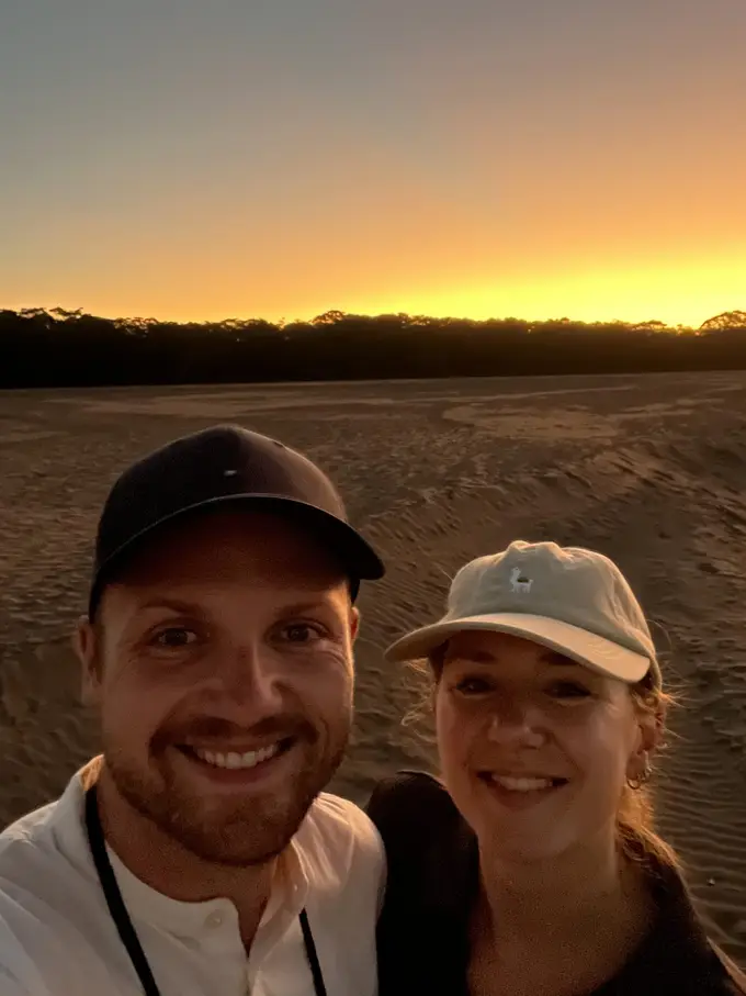 Selfie von Max und Rebecca auf einer Sandbank, hinter ihnen ein orange leuchtender Streifen über dem Regenwald am Horizont