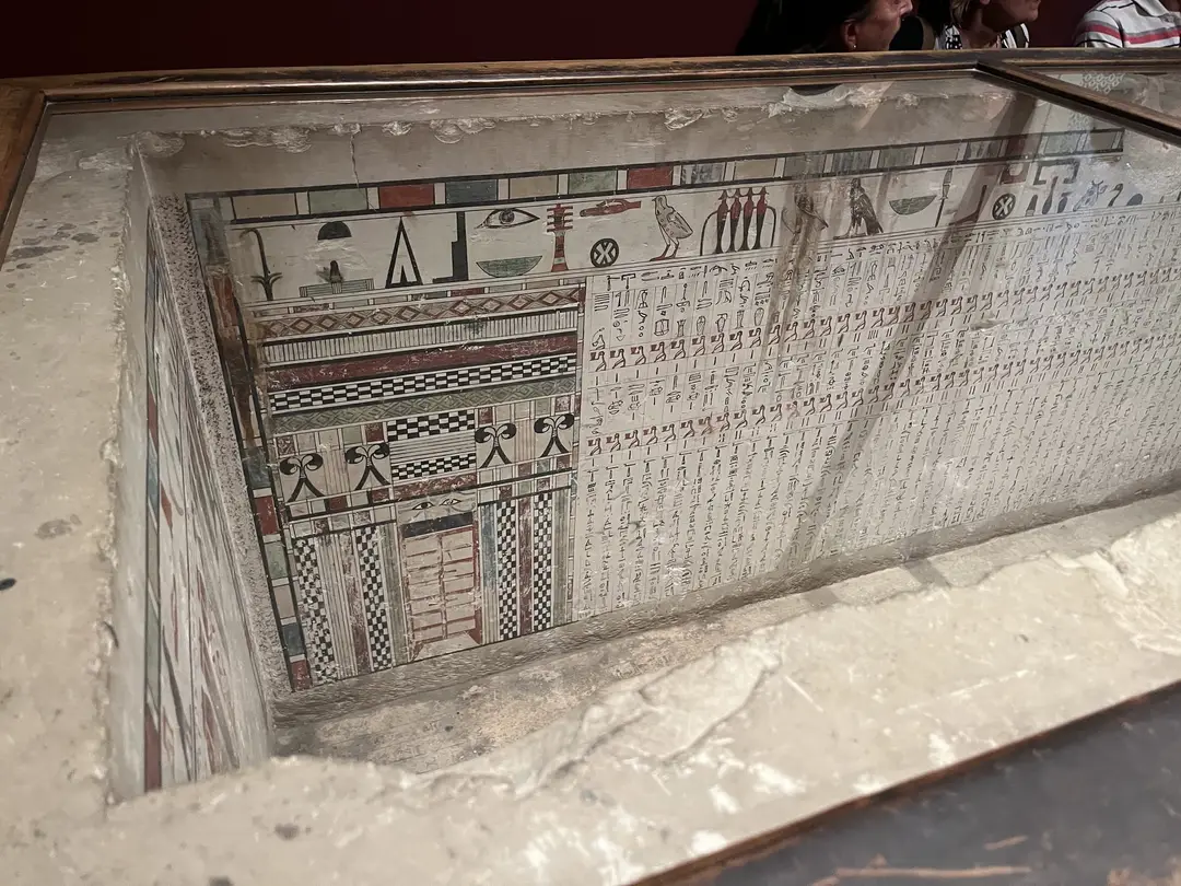 Blick durch eine Glasabdeckung in einen offenen Steinsarkophag, die Innenwände bemalt mit Hieroglyphenreihen, farbigen Mustern und einer Scheintür