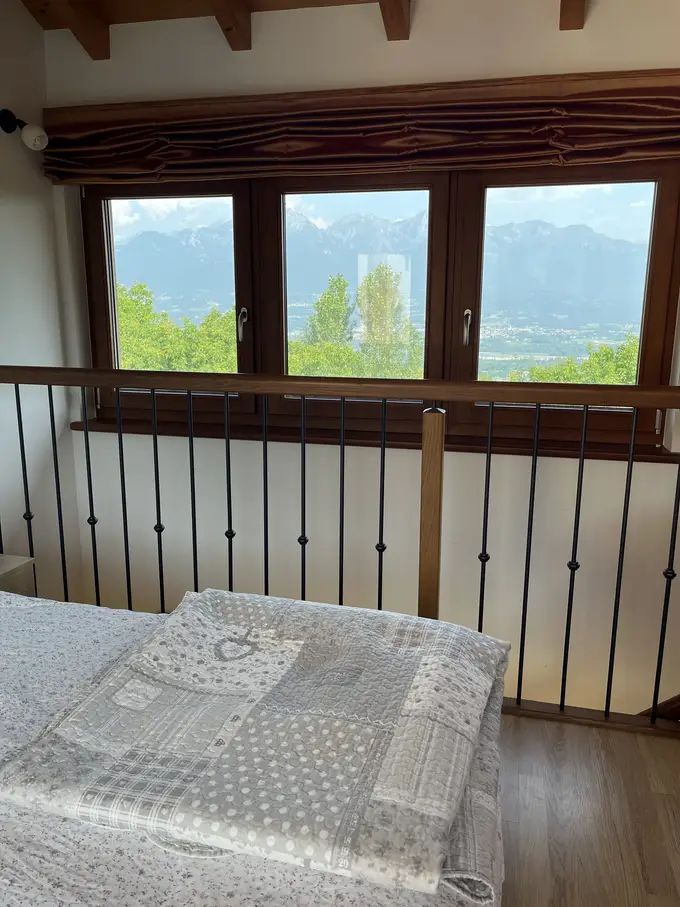 Ein Bett unter der Dachschräge mit Holzbalken, dahinter ein Geländer und drei Fenster mit Blick über Bäume auf ein Tal und eine Bergkette
