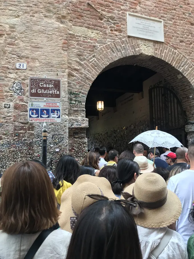 Menschenmenge mit Sonnenhüten und Schirmen vor dem Torbogen mit dem Schild Casa di Giulietta