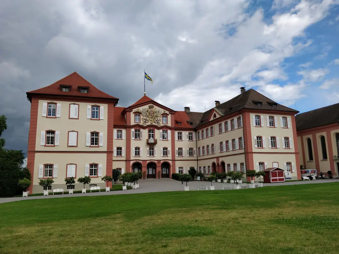 Das barocke Schloss auf der Mainau mit hell-roter Fassade, Wappen über dem Portal und einer Fahne auf dem Dach, davor eine große Rasenfläche