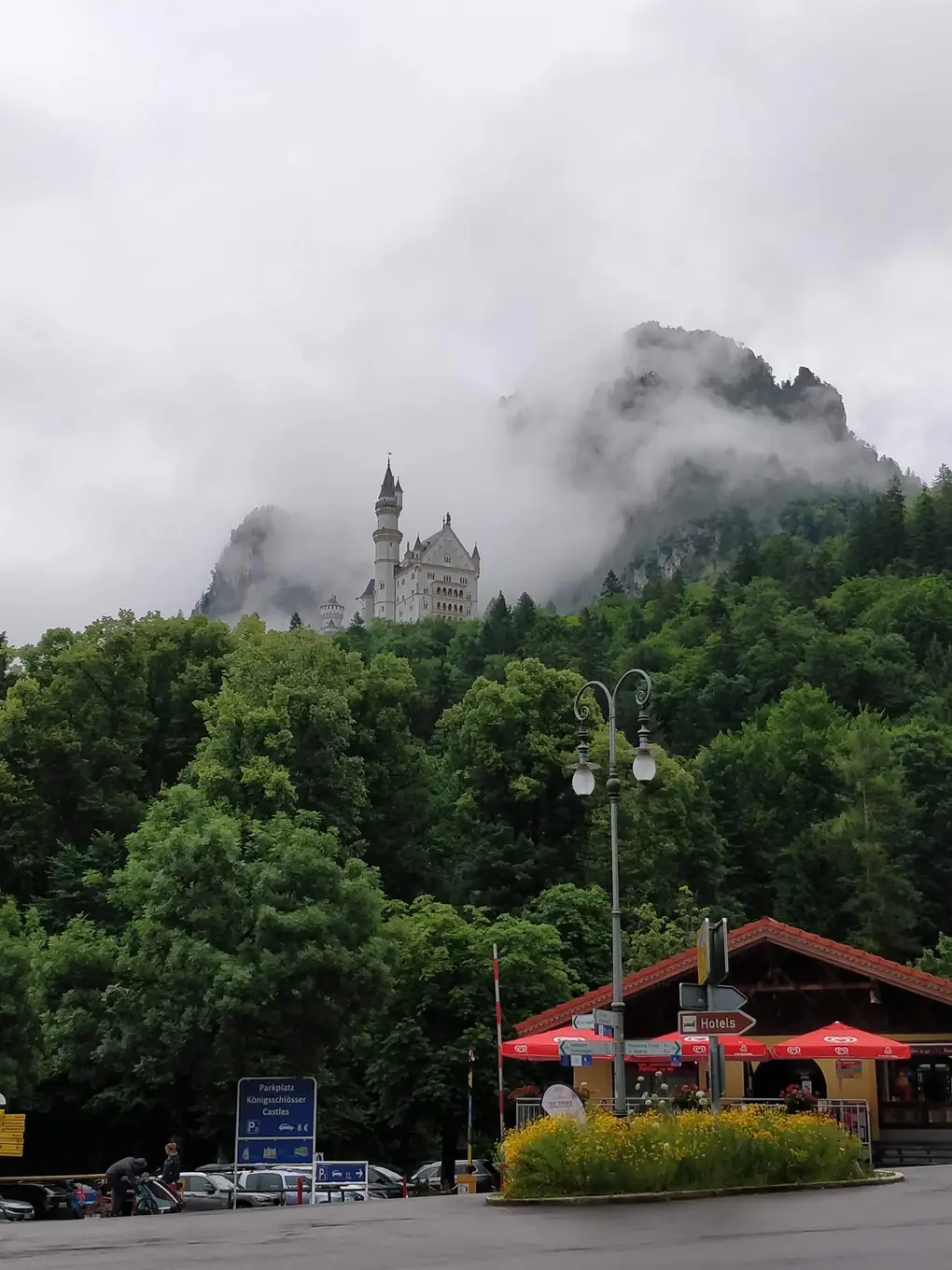 Neuschwanstein klein und weiß hoch über einem bewaldeten Hang im Nebel, unten ein Parkplatzschild, Wegweiser und ein Kiosk mit roten Sonnenschirmen