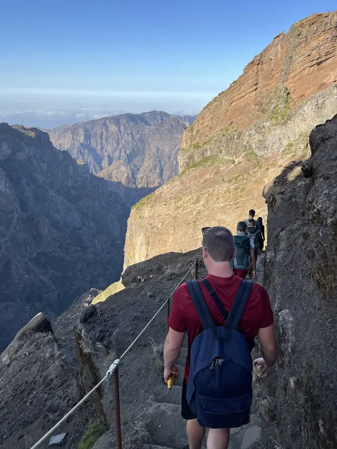 Ein Wanderer mit Rucksack auf einem schmalen Pfad an einer steilen, rötlichen Felswand