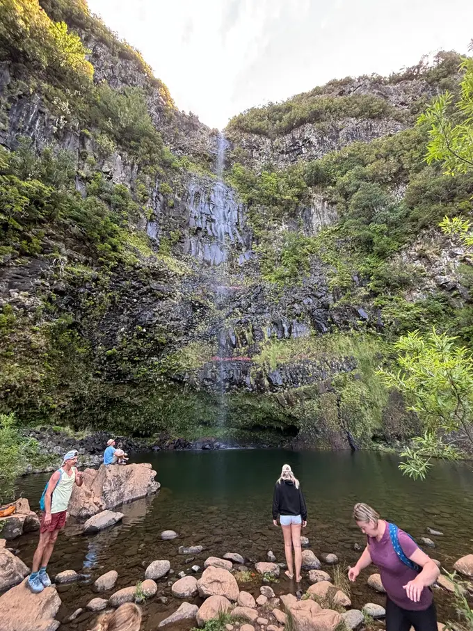 Ein schmaler Wasserfall stürzt in ein dunkles Becken, drei Personen stehen am felsigen Ufer