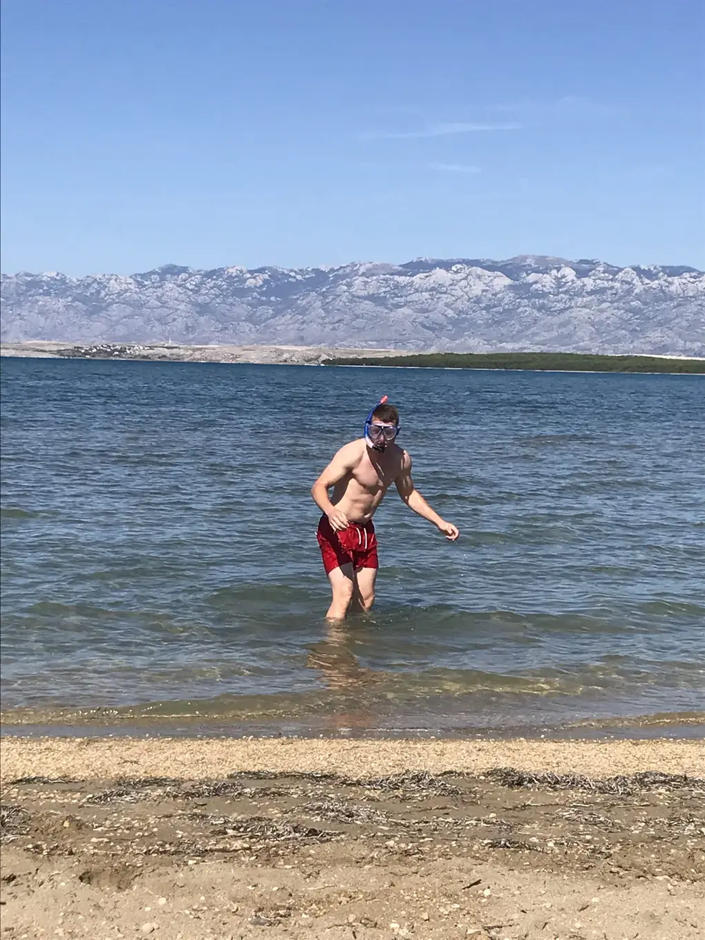 Ein Mann in roter Badehose mit Taucherbrille und Schnorchel steht im flachen Wasser der Lagune, dahinter ein Gebirgszug