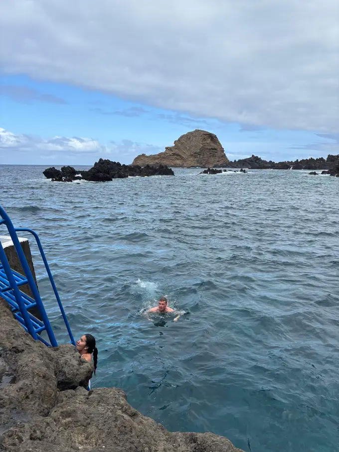 Zwei Personen schwimmen im Wasser neben einer blauen Leiter am Fels, eine weitere Person sitzt am Ufer