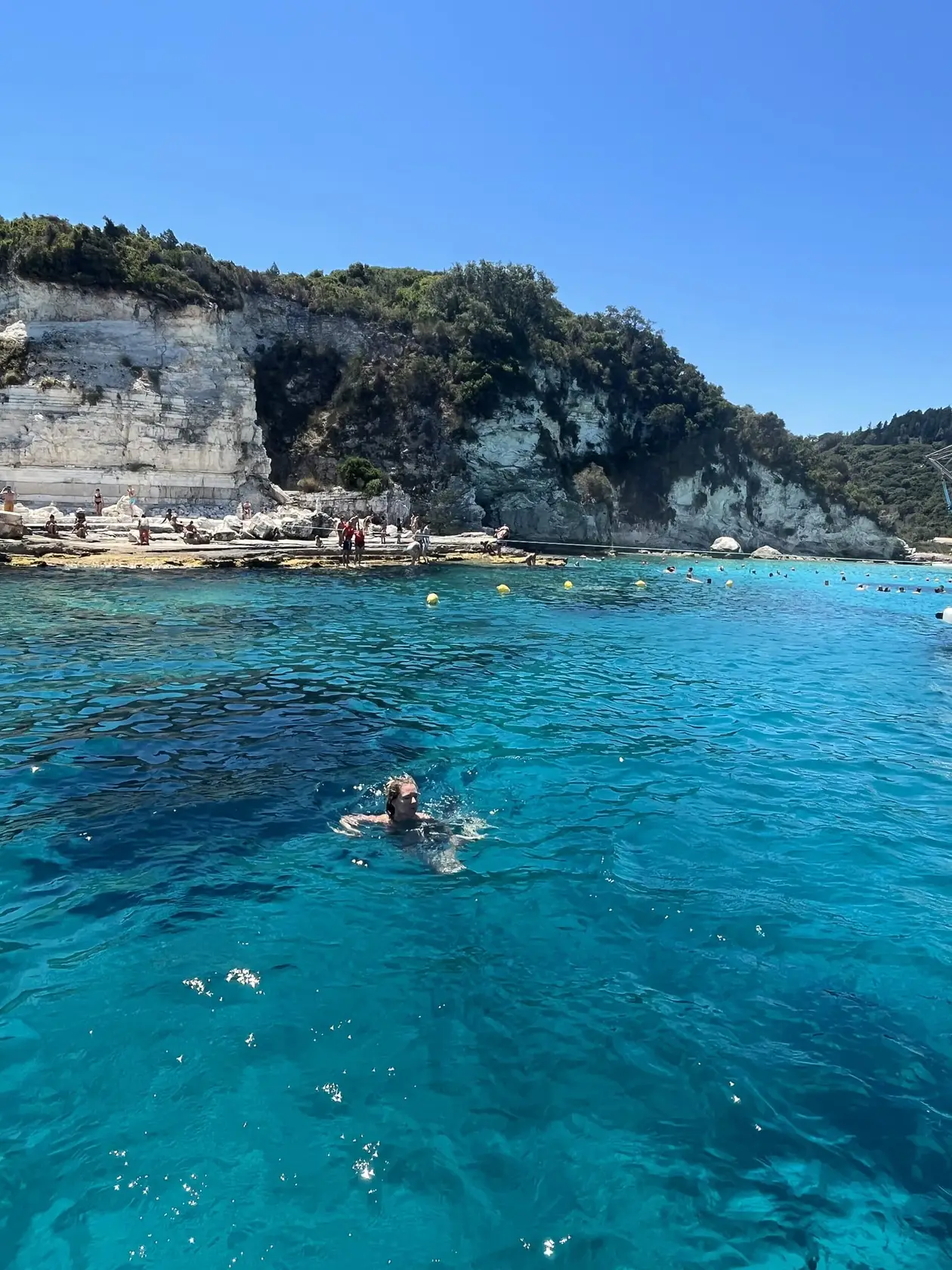 Ein Schwimmer in türkisem Wasser über hellem Grund, dahinter eine helle Felsküste mit bewaldetem Hang
