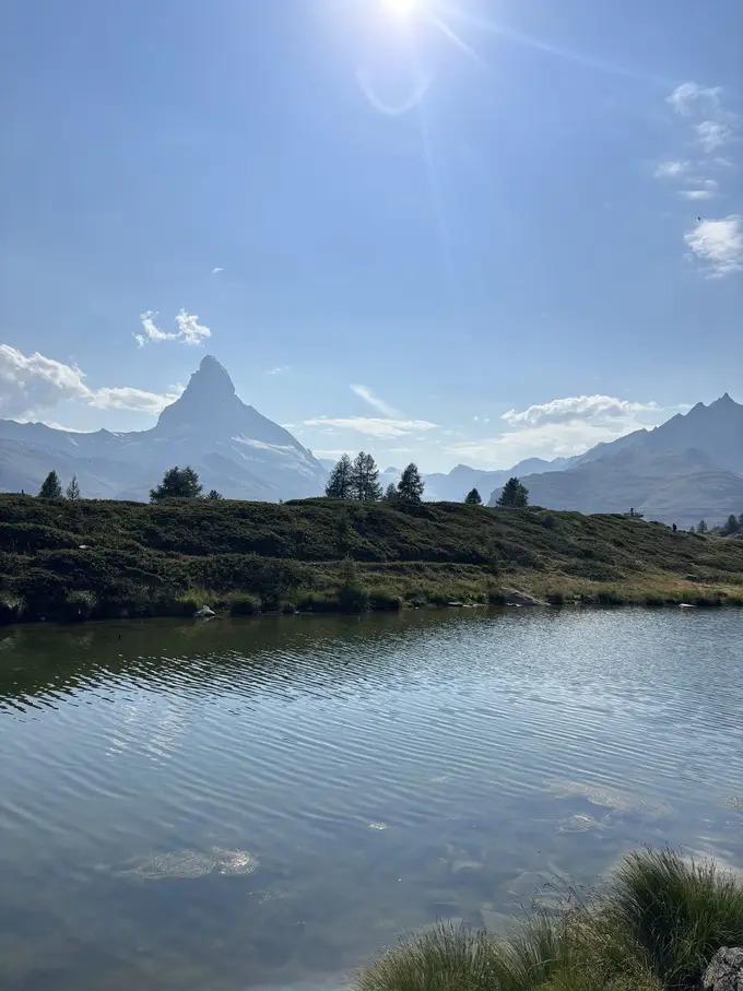 Ein kleiner Bergsee vor einem Hügel mit Nadelbäumen, dahinter das Matterhorn und die Sonne am Himmel