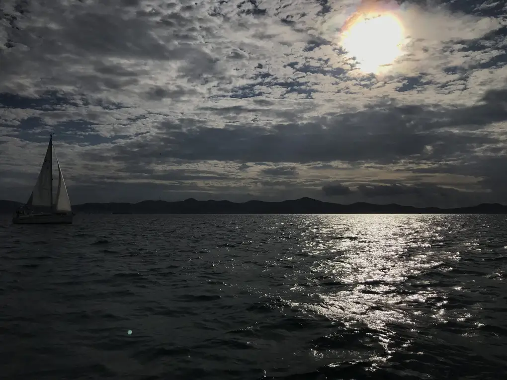 Ein Segelboot im Gegenlicht auf glitzerndem Wasser, die Sonne hinter Wolken, am Horizont Inseln