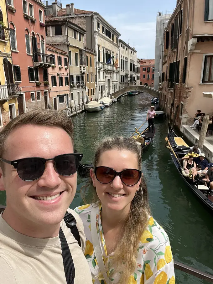 Selfie von Max und Rebecca auf einer Brücke, hinter ihnen ein schmaler Kanal zwischen bunten Häusern mit Gondeln und einer Steinbrücke