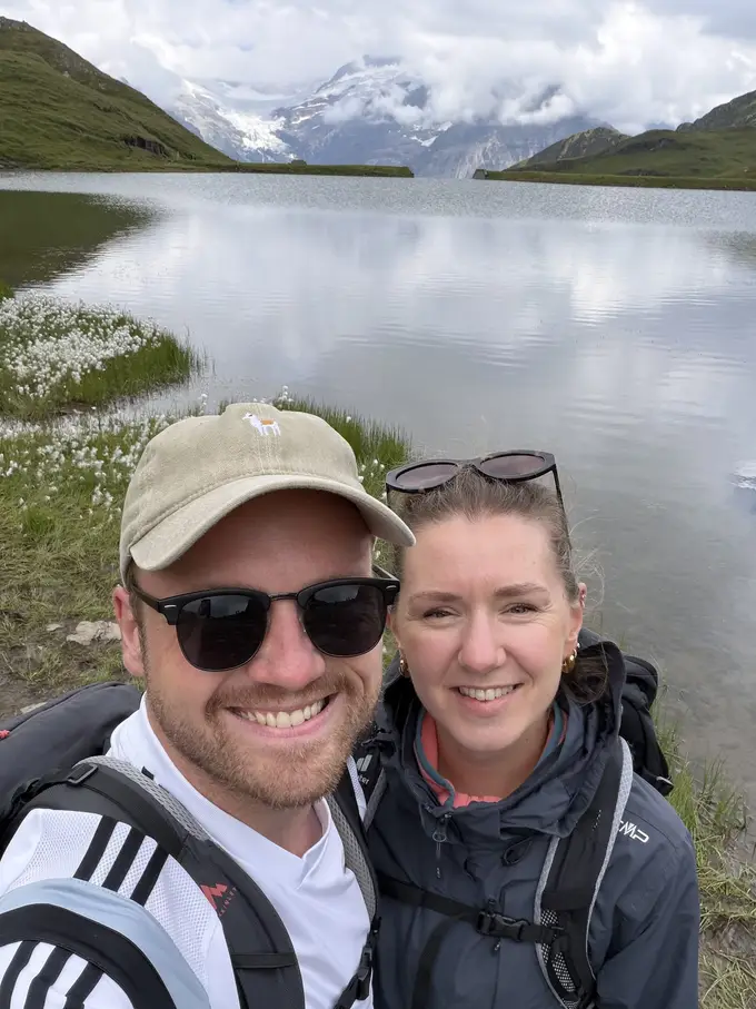 Selfie von Max und Rebecca am Ufer des Bachalpsees, hinter ihnen Wollgras, das Wasser und ein Gletscher in den Wolken
