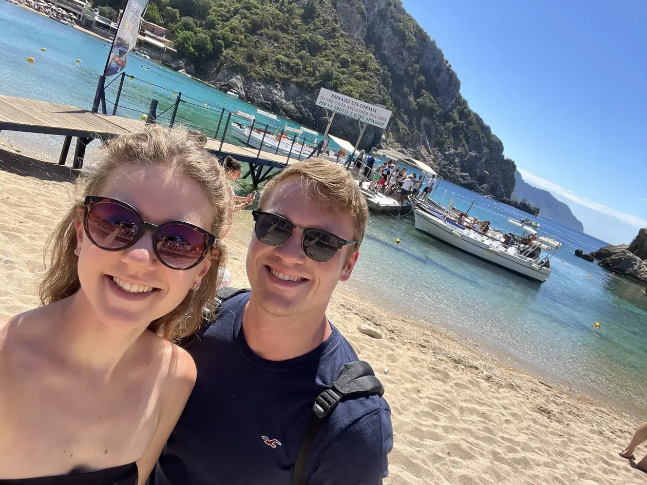 Selfie von Rebecca und Max am Sandstrand, hinter ihnen ein Bootssteg, Ausflugsboote und ein bewaldeter Berg