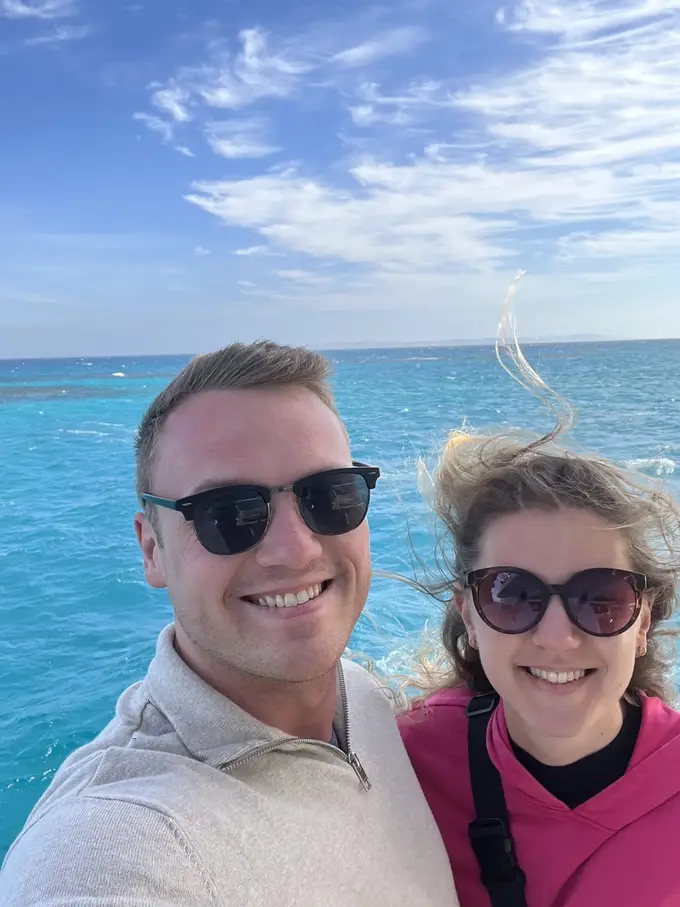 Selfie von Max und Rebecca an Deck, Rebeccas Haare wehen im Wind, dahinter türkises Wasser und die Küste
