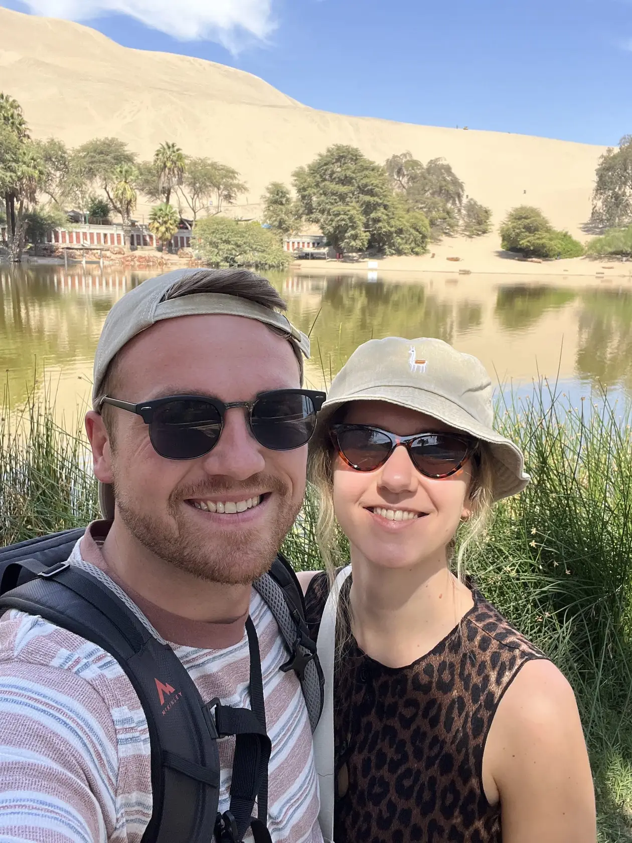 Max und Rebecca mit Sonnenbrillen und Hüten am Ufer der Lagune, dahinter Palmen und eine hohe Sanddüne
