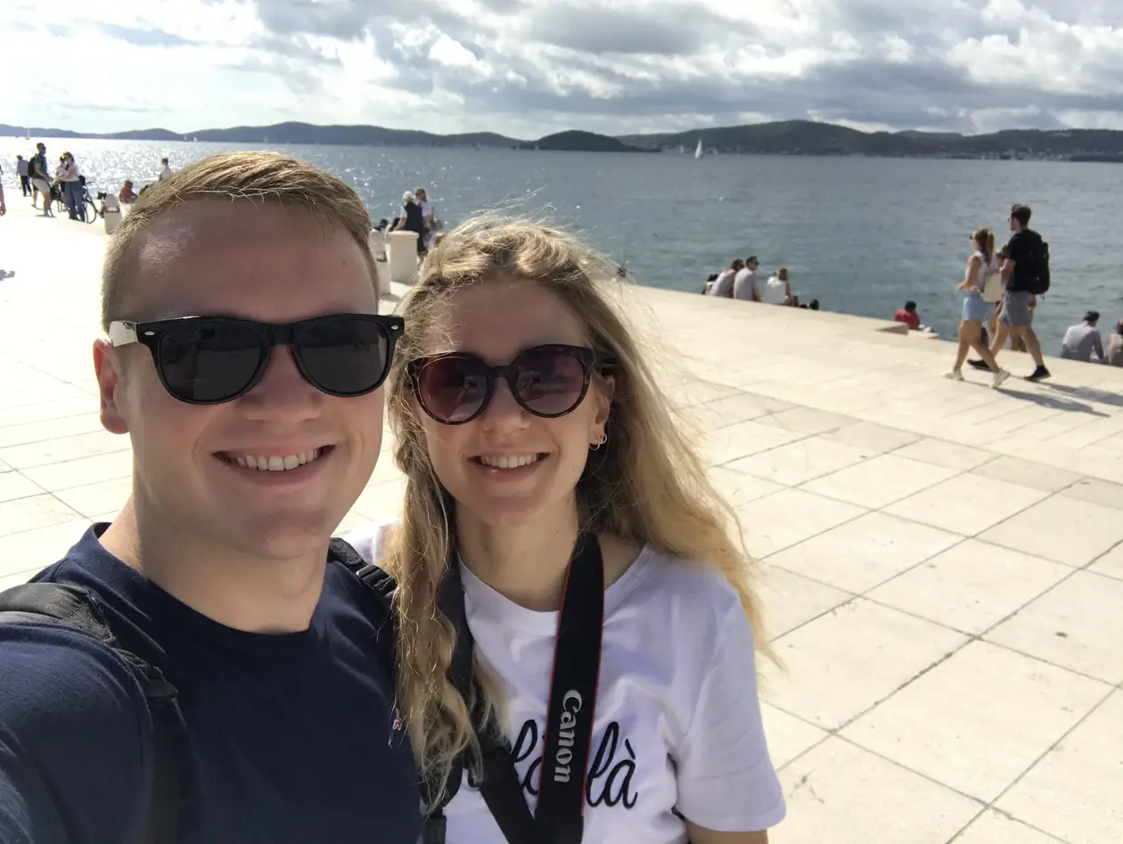 Selfie von Max und Rebecca mit Sonnenbrillen an der Uferpromenade, hinter ihnen Menschen auf hellen Steinstufen, das Meer und flache Inseln