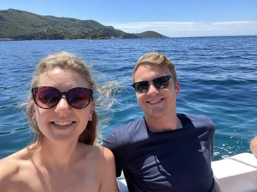Selfie von Rebecca und Max mit Sonnenbrillen auf einem Boot, hinter ihnen offenes Meer und eine bewaldete Küste