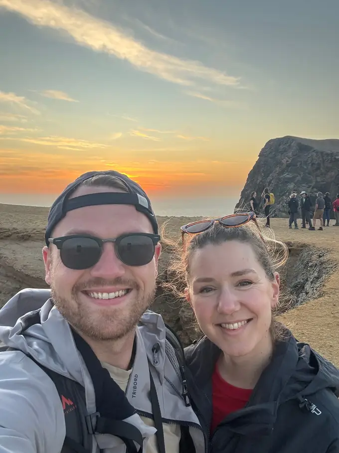 Max und Rebecca machen ein Selfie auf der Klippe, hinter ihnen geht die Sonne orange über dem Meer unter