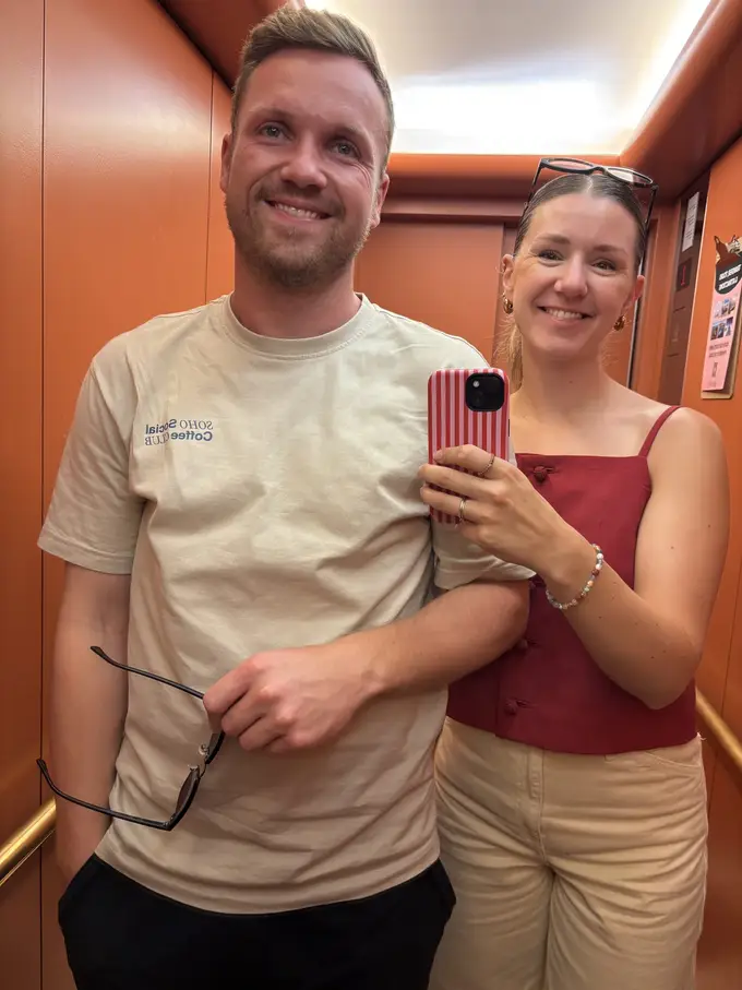Max und Rebecca machen ein Spiegelselfie in einem Aufzug mit orangefarbenen Wänden